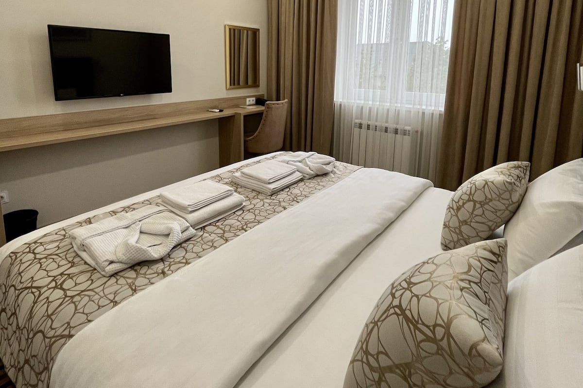 A hotel room with a bed, a TV mounted on the wall, a window with curtains, and a bedside table.