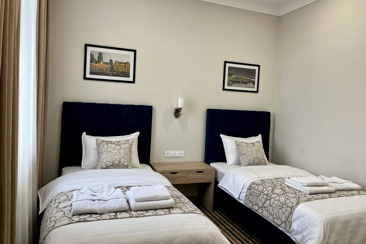 A hotel room with two beds, each with a dark headboard, white bedding, and a patterned throw. There are two framed pictures on the wall, a bedside table with a lamp, and a window with curtains.