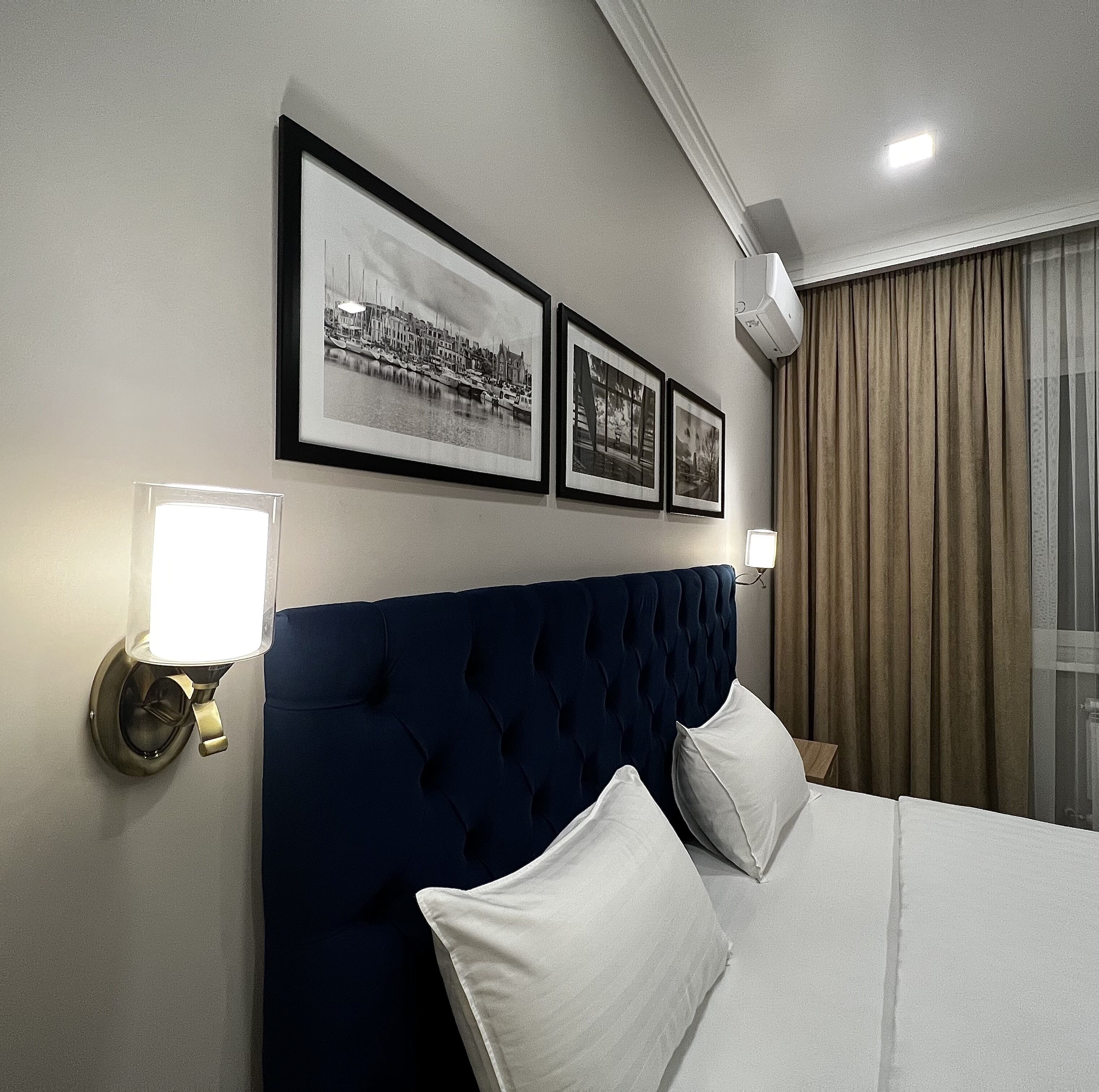 A hotel room with a large bed, a headboard, two wall-mounted picture frames, a wall-mounted lamp, and a window with curtains.