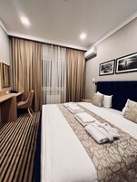 Habitación de hotel con una cama grande, un escritorio, una silla y vistas de la ciudad a través de cortinas translúcidas.