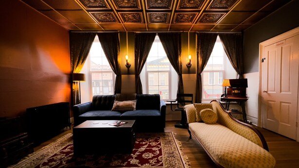 Ein gemütliches Wohnzimmer mit einer Couch, einem gemusterten Teppich und einem Couchtisch. Drei Fenster mit Vorhängen, eine Lampe und eine Tür mit der Aufschrift 'PRIVATE'.