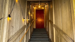 Eine Holztreppe mit einer roten Tür oben, umgeben von Wänden mit freiliegenden Holzbeschlägen und Lichterketten.