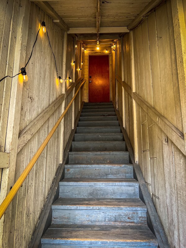 Un escalier en bois, avec une porte rouge en haut, entouré de murs aux panneaux de bois apparents et illuminé de guirlandes lumineuses.