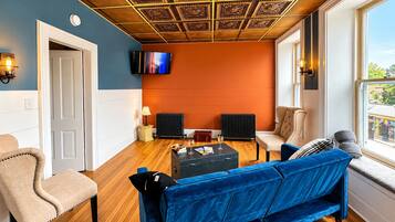 Un salon avec un canapé bleu, un fauteuil beige et un canapé en velours bleu. On y trouve un téléviseur fixé au mur, une lampe et un radiateur.
