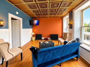 A living room with a blue sofa, a beige armchair, and a blue velvet sofa. There is a television mounted on the wall, a lamp, and a radiator.
