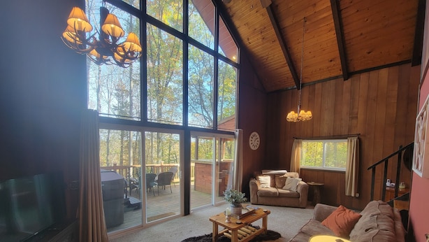 A spacious living room with a large window, a wooden ceiling, a sofa, a coffee table, and a television. - Hocking Hills secluded Mayapple Cabin - 8 miles from Cantwell Cliffs! ~Hot Tub~ (Rockbridge)