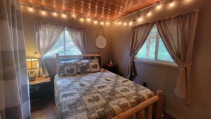 A cozy bedroom with a wooden bed frame, a quilt with animal patterns, two windows with curtains, a bedside table with a lamp, and string lights on the ceiling. - Hocking Hills secluded Mayapple Cabin - 8 miles from Cantwell Cliffs! ~Hot Tub~ (Rockbridge)