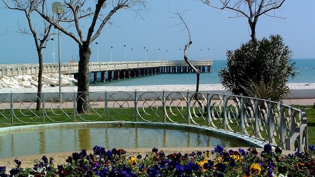 A waterfront promenade with a curved metal railing, a bridge, and a row of trees.
