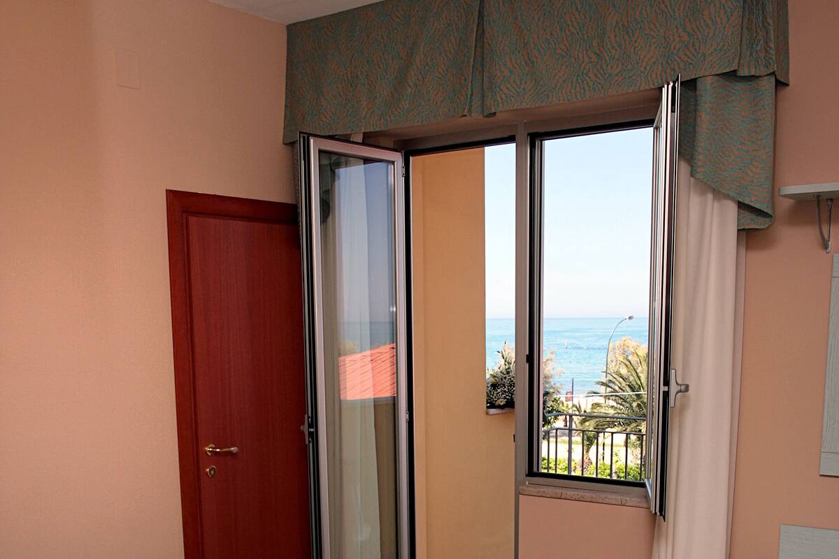 A bedroom with a bed, a door, a sliding glass door leading to a balcony, and a view of the sea.
