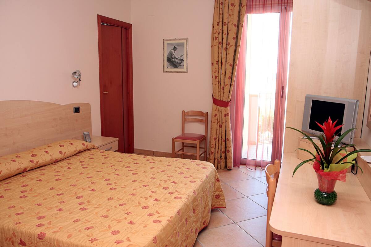 A bedroom with a bed, a chair, a desk, a television, and a window with curtains.