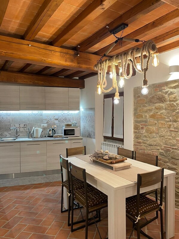 Een moderne keuken met een accentmuur van steen, houten balken in het plafond, en een eethoek met een rustieke kroonluchter.