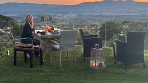A dining table with chairs, a bottle, glasses, and a candle on a grassy hill overlooking a valley. - Villa Borgo Soale in Tuscany—Reggello. Private pool and Wi-Fi (Toscana)