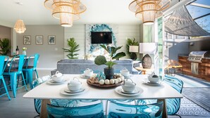 A modern dining area with a white table set for tea service, surrounded by blue chairs, and a large window offering a view of the outdoors. - Disney-Themed Home with Pool and Game Room (Kissimmee)