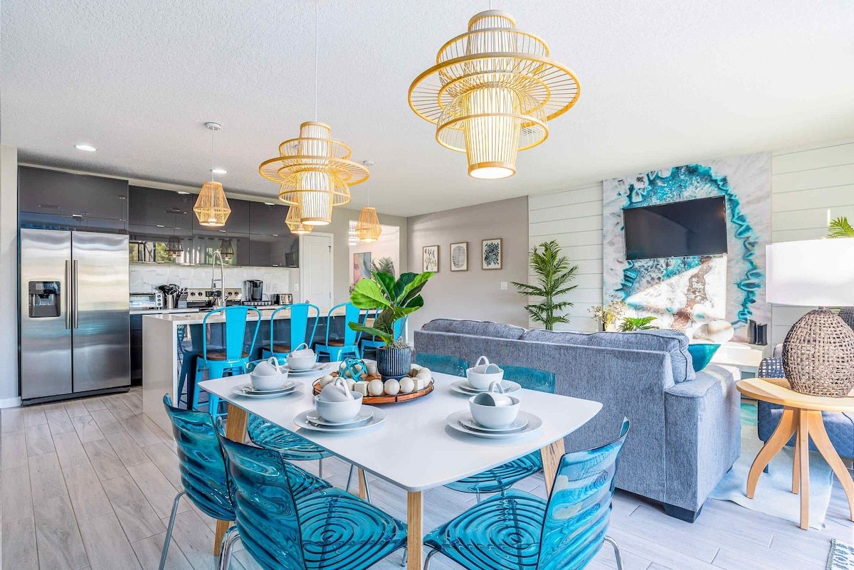 A modern kitchen with stainless steel appliances, a dining area with blue chairs, and a living room with a grey sofa.