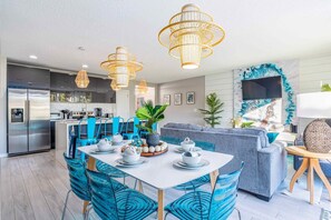 A modern kitchen with stainless steel appliances, a dining area with blue chairs, and a living room with a grey sofa.