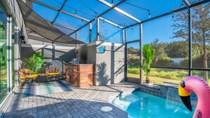 An indoor swimming pool with a glass enclosure, a wooden bench, and a pink flamingo decoration. - Disney-Themed Home with Pool and Game Room (Kissimmee)