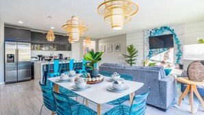 A modern kitchen with stainless steel appliances, a dining area with blue chairs and a white table set for four, and a living area with a grey sofa and a TV mounted on the wall. - Disney-Themed Home with Pool and Game Room (Kissimmee)