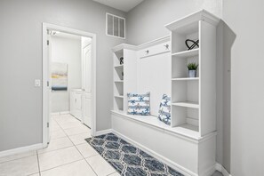 A hallway with a white door, a white wall, a white shelf unit with decorative pillows, and a patterned rug. - Sleeps 21 / Disney & Universal Retreat w/ Game Room + Pool, Spa (Kissimmee)