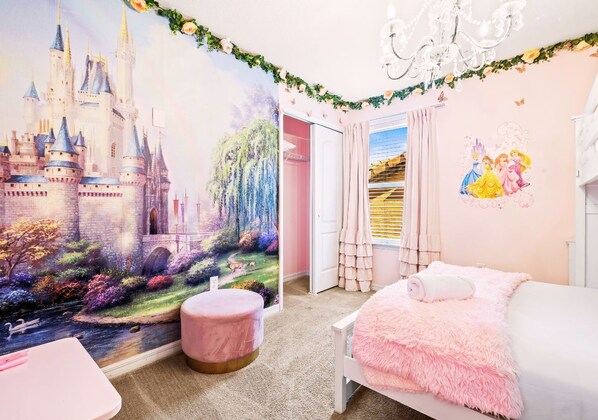 Une chambre avec une fresque représentant un château, un pouf rose, une couverture rose et moelleuse, et un lustre.