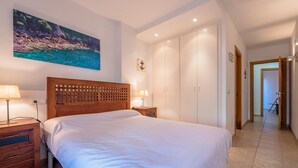 Een slaapkamer met een groot bed, houten hoofdbord, nachtkastjes met lampen, een kledingkast en een schilderij van een kustlandschap.