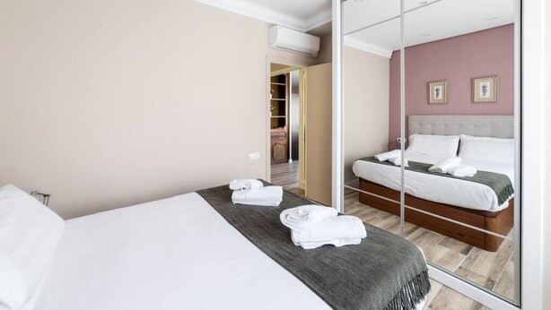 Une chambre d’hôtel avec un grand miroir, un lit recouvert de draps blancs, un plaid gris et une tête de lit en bois.