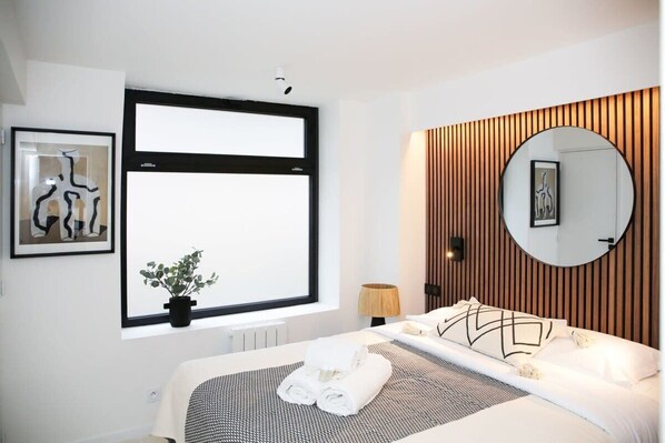 Una habitación de hotel moderna con un ventanal grande, una cama con almohada de estampado geométrico, un espejo redondo y una lámpara fijada a la pared.