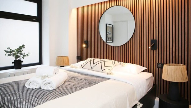 A modern hotel room with a bed, a round mirror, a potted plant, and a nightstand with a lamp.