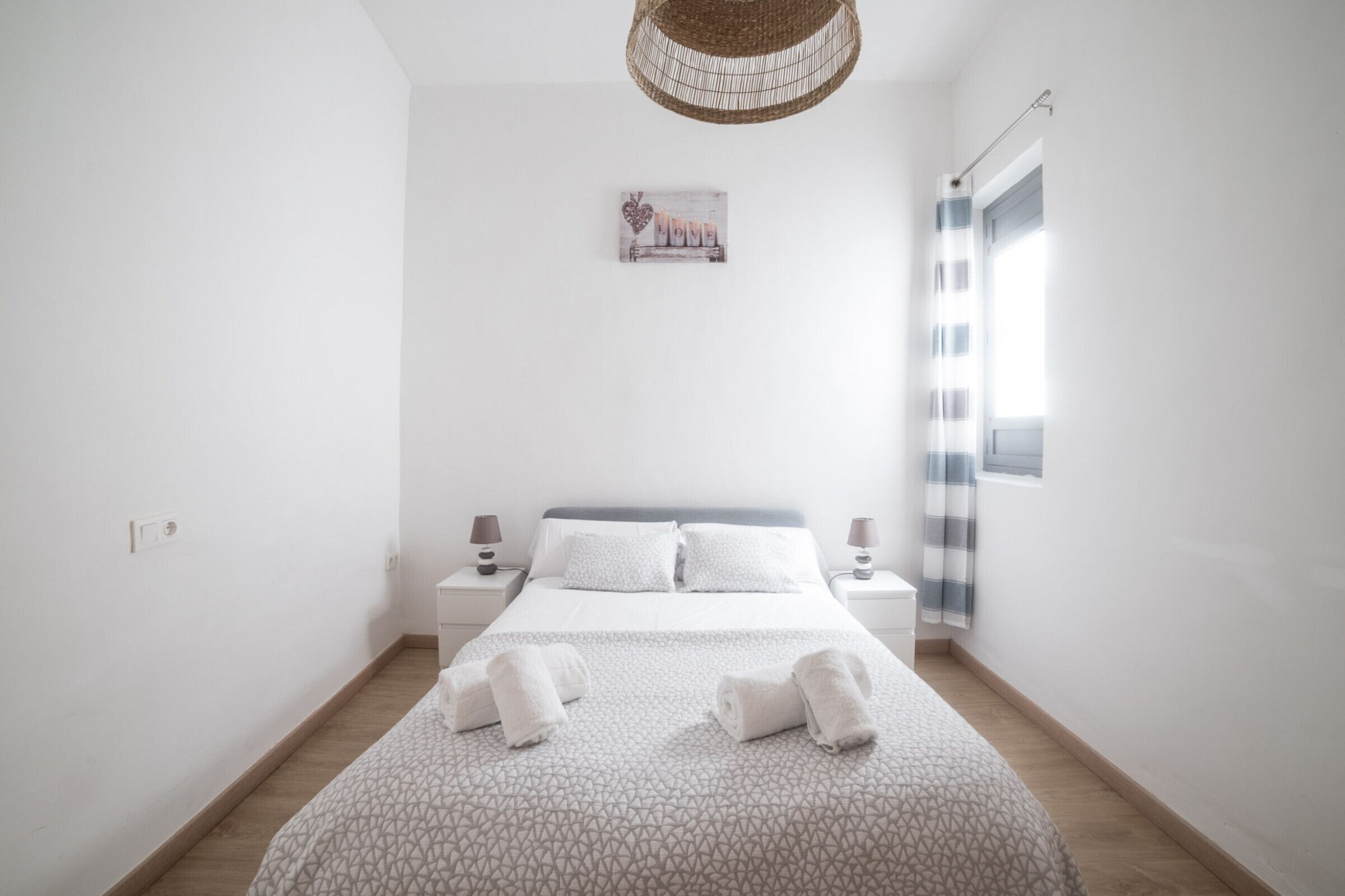 Um quarto arrumado com uma cama, duas mesas de cabeceira, um candeeiro pendente e uma janela com cortinas.