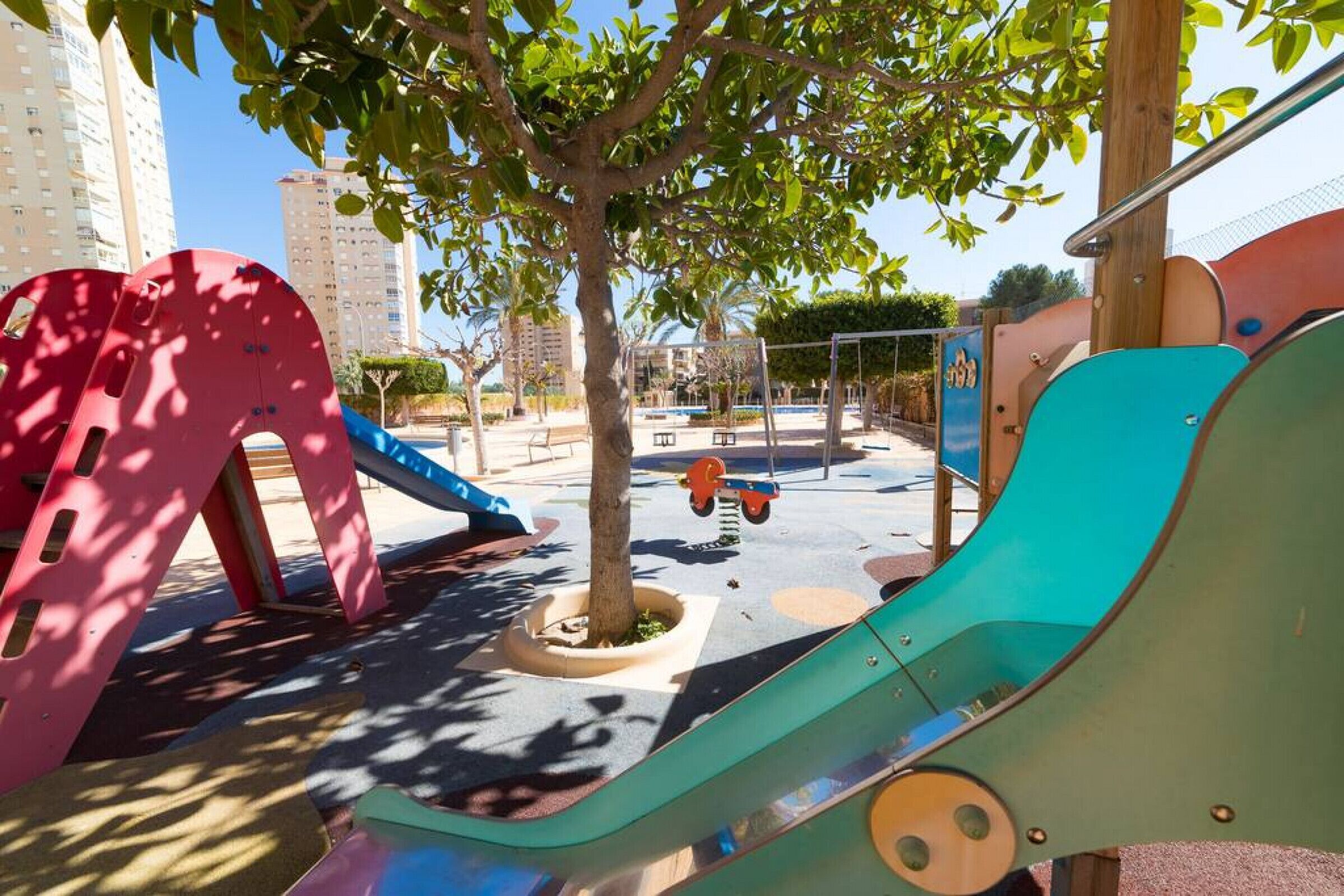 Un parque infantil con toboganes y columpios de colores, rodeado de árboles y edificios.
