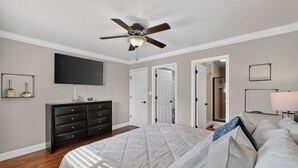 Um quarto com uma cama, cômoda, ventilador de teto, televisão e uma porta que dá para outro ambiente.