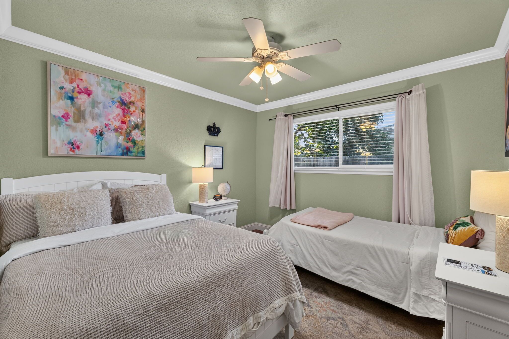 A bedroom with two beds, a ceiling fan, a window with curtains, and a painting on the wall.