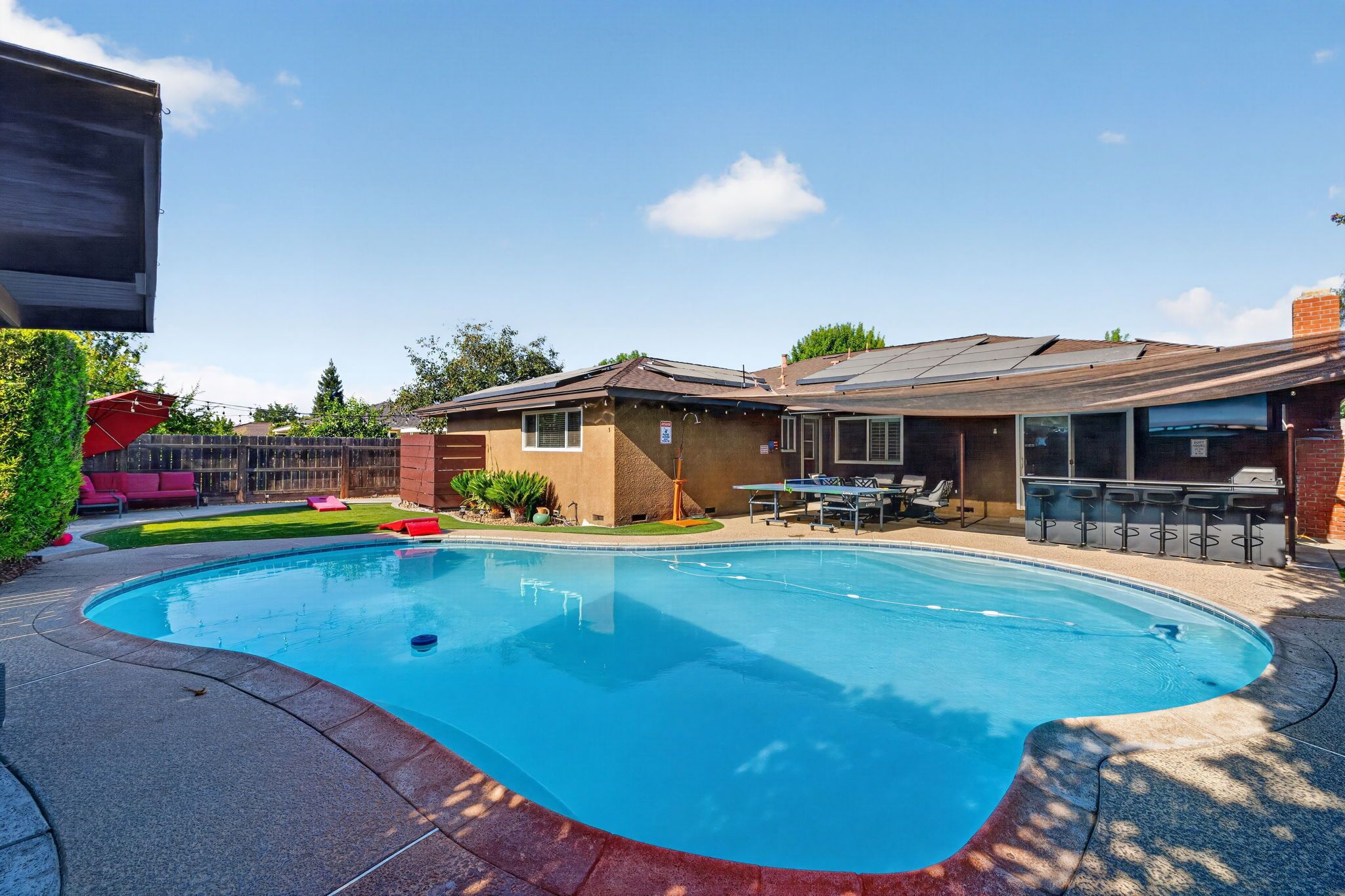Une piscine dont le centre est plus profond que le reste, entourée d’un patio aménagé avec des meubles d’extérieur et un espace salon couvert.