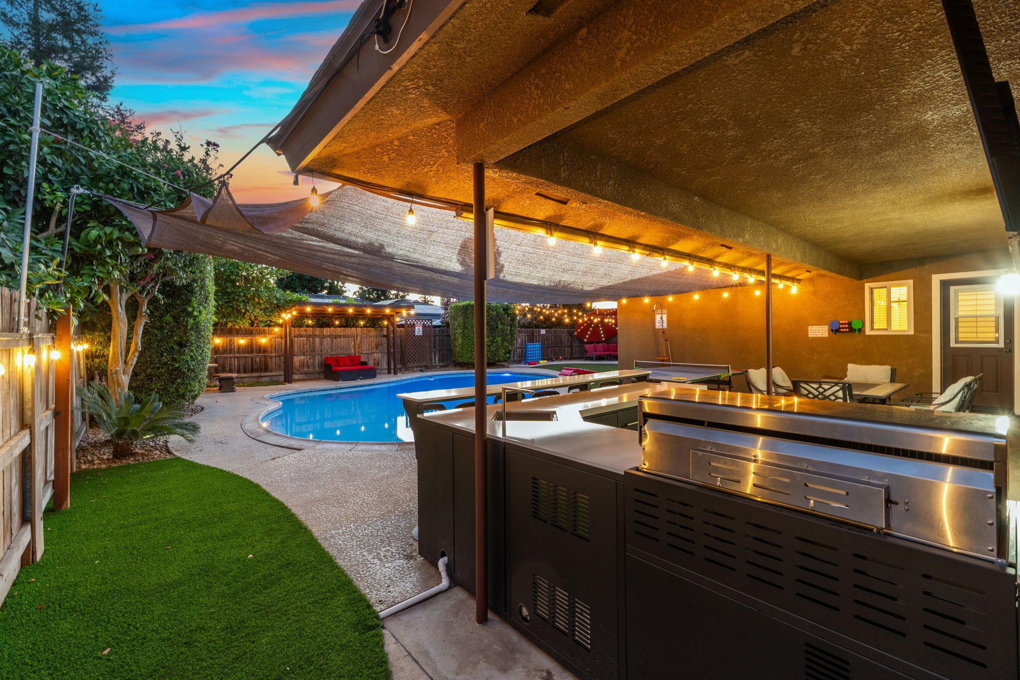 Un área de piscina al aire libre con una estación de parrillada, luces colgantes y una zona de estar cubierta.