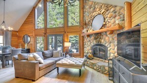A cozy living room with a stone fireplace, wooden walls, and a large window overlooking a forest. - Luxe Cabin in the Woods: Walk to Lake! (Gaston)