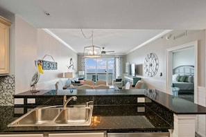 A kitchen with a black granite countertop, stainless steel sink, and a dining area with a view of the sky.