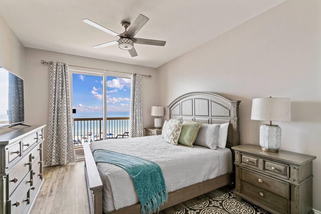 A bedroom with a bed, a ceiling fan, a nightstand, a dresser, and a view of the ocean.