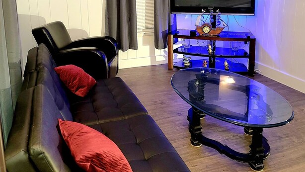 Ett vardagsrum med en svart lädersoffa, röda kuddar, ett soffbord i glas, en platt-TV och en tavla på väggen.