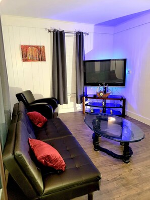 Un salon avec un canapé en cuir noir, des coussins rouges, une table basse en verre, un téléviseur à écran plat et un tableau accroché au mur.