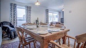 A dining area with a wooden table set for four, two chairs on each side, and a view of the sea through large windows.