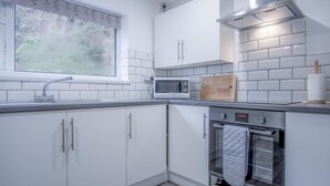 A modern kitchen with white cabinets, a microwave, an oven, a window with a view of greenery, and a wooden cutting board.