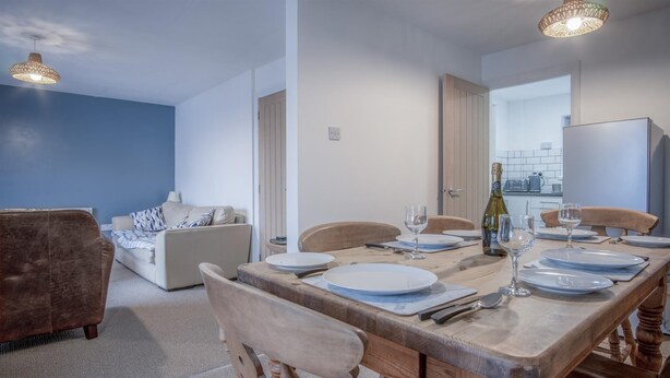 A dining area with a wooden table set for four, a bottle of wine, and a view into the kitchen.