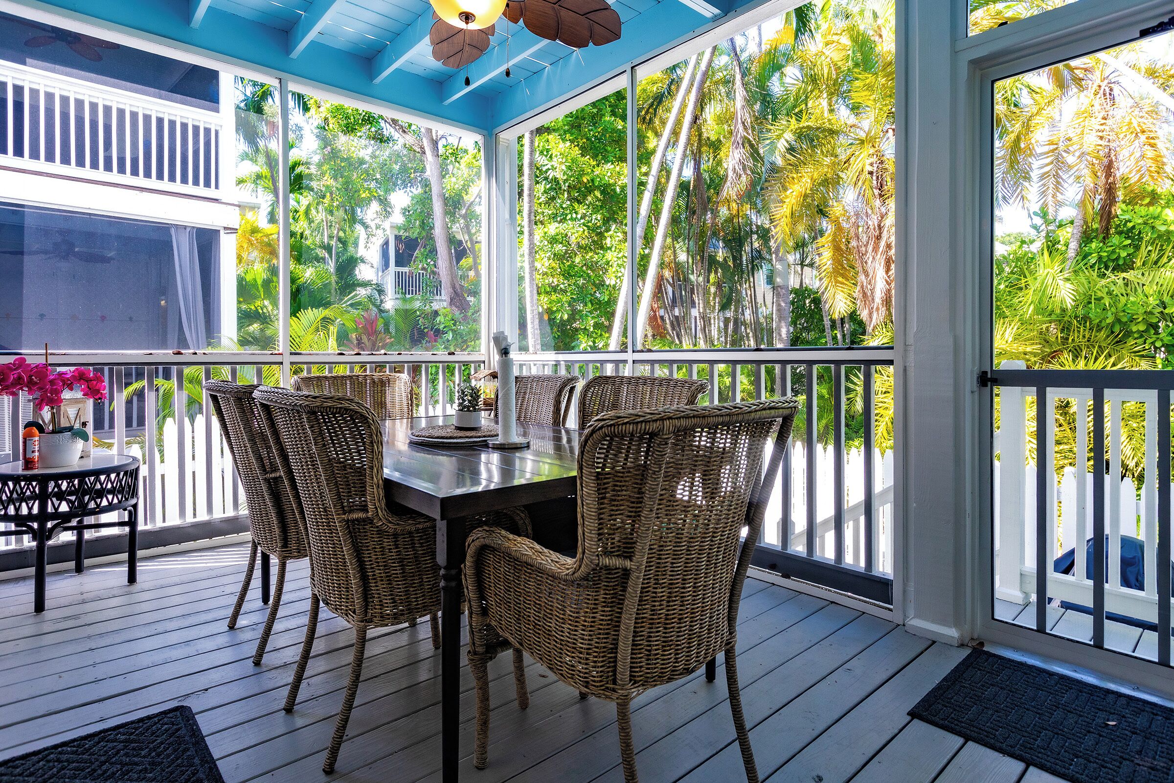 A covered outdoor patio with a dining table and wicker chairs, surrounded by large windows offering views of tropical vegetation.