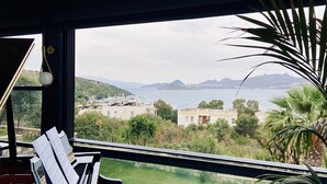 A piano with sheet music, a glass table, and a potted plant in a room with a view of a marina and mountains. - Roas Hotel (Bodrum)