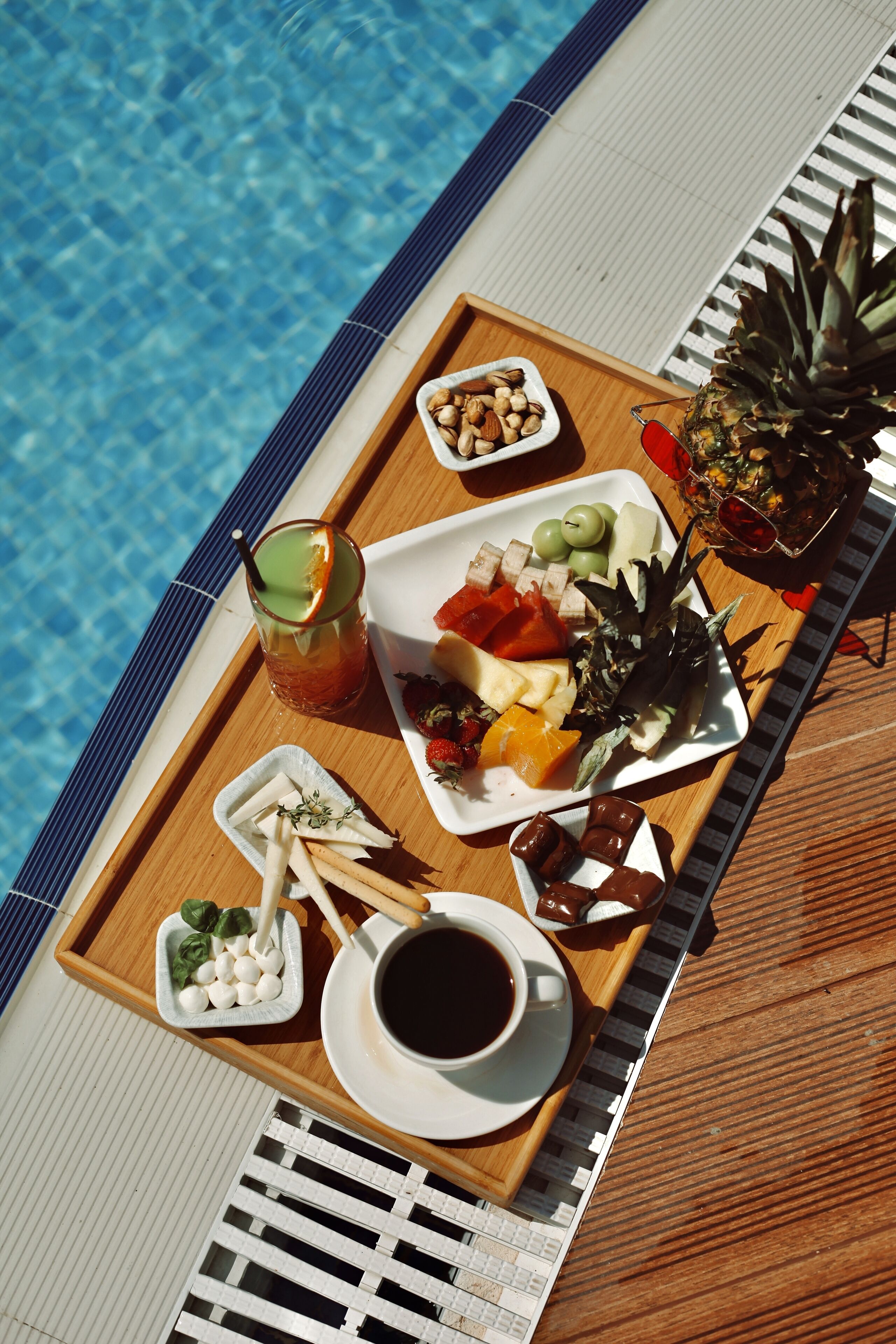 A wooden tray with a variety of food items and a drink by a poolside.