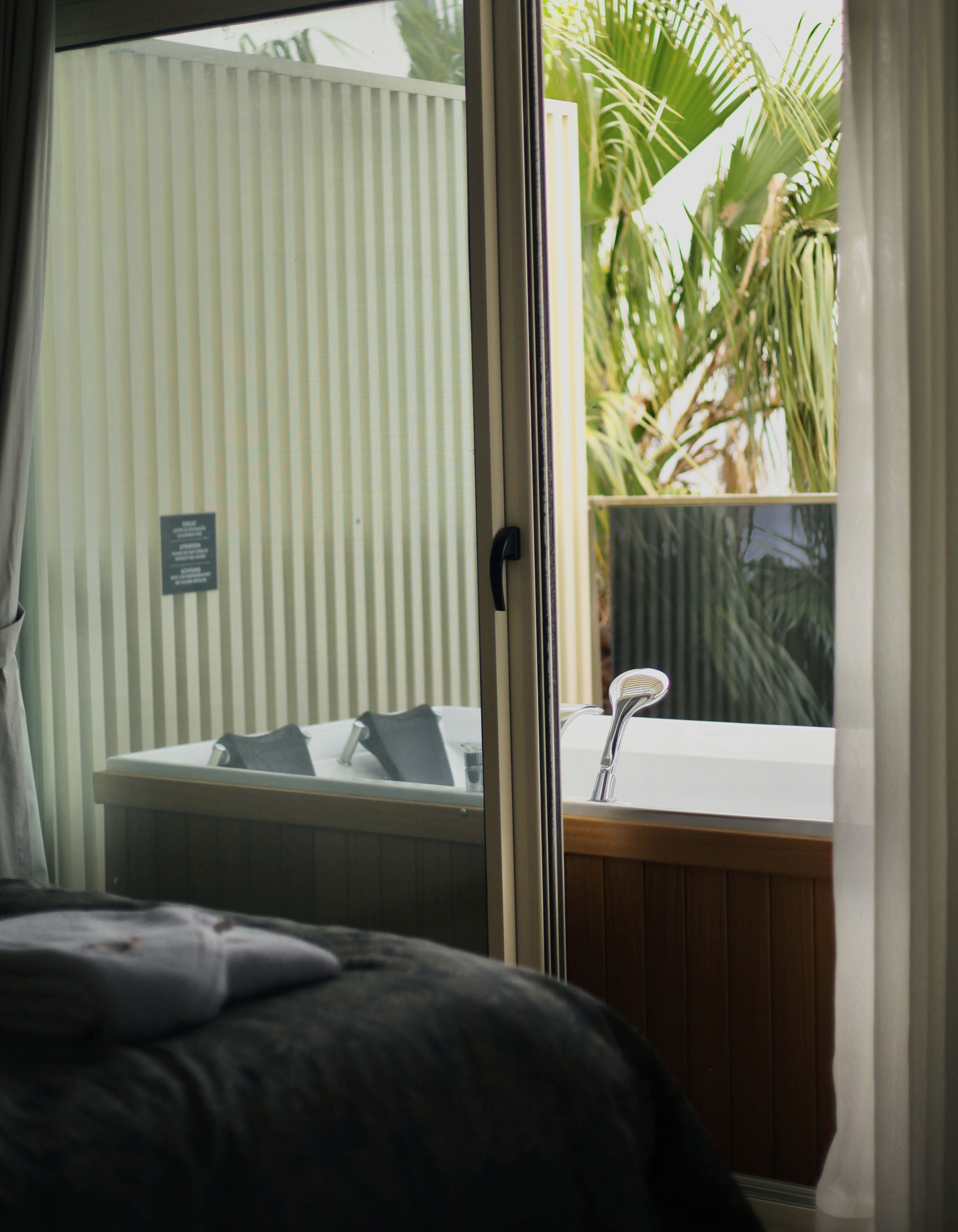 Una habitación con cama, ventana con persianas y vista a palmeras, además de un lavamanos pequeño.