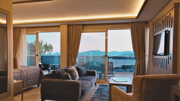 A hotel room with a bed, a sofa, a chair, a TV, a table, and large windows with a view of the sea and mountains. - Roas Hotel (Bodrum)