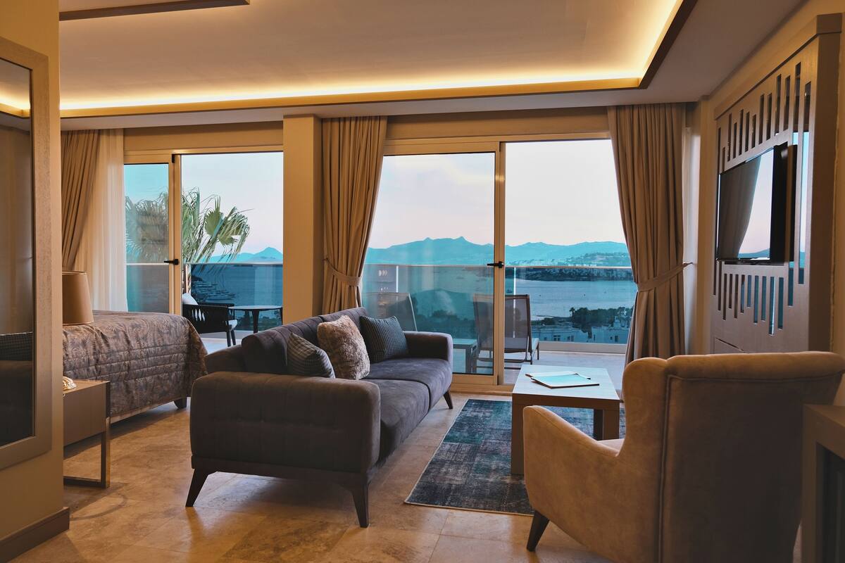 Quarto de hotel com cama, sofá, poltrona, televisão, mesa e grandes janelas com vista para o mar e montanhas.