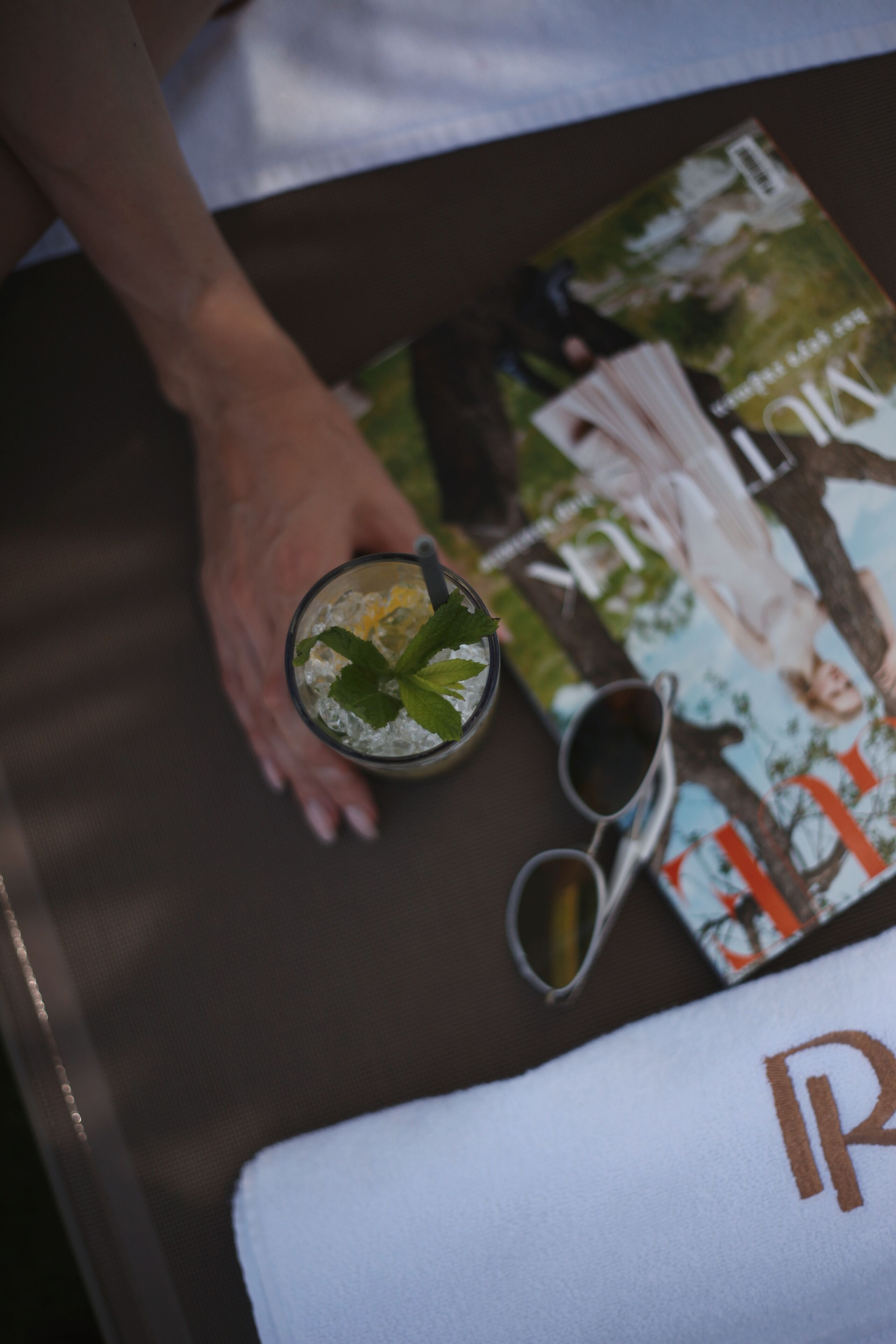 A table with a magazine, a glass of iced beverage with mint, and a pair of sunglasses.