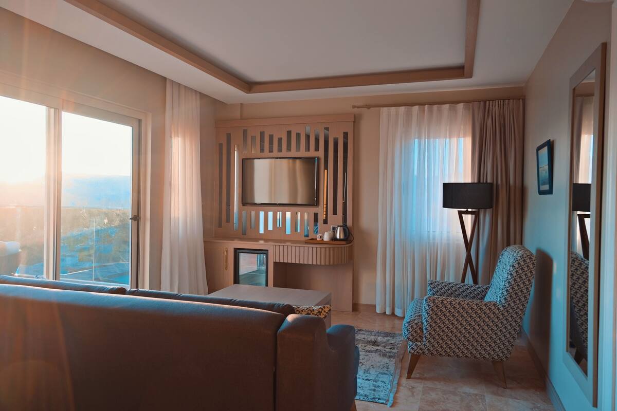 Quarto de hotel com uma grande janela, televisão de tela plana presa a um painel de madeira, sofá, poltrona estampada, escrivaninha com abajur e vista para o exterior.