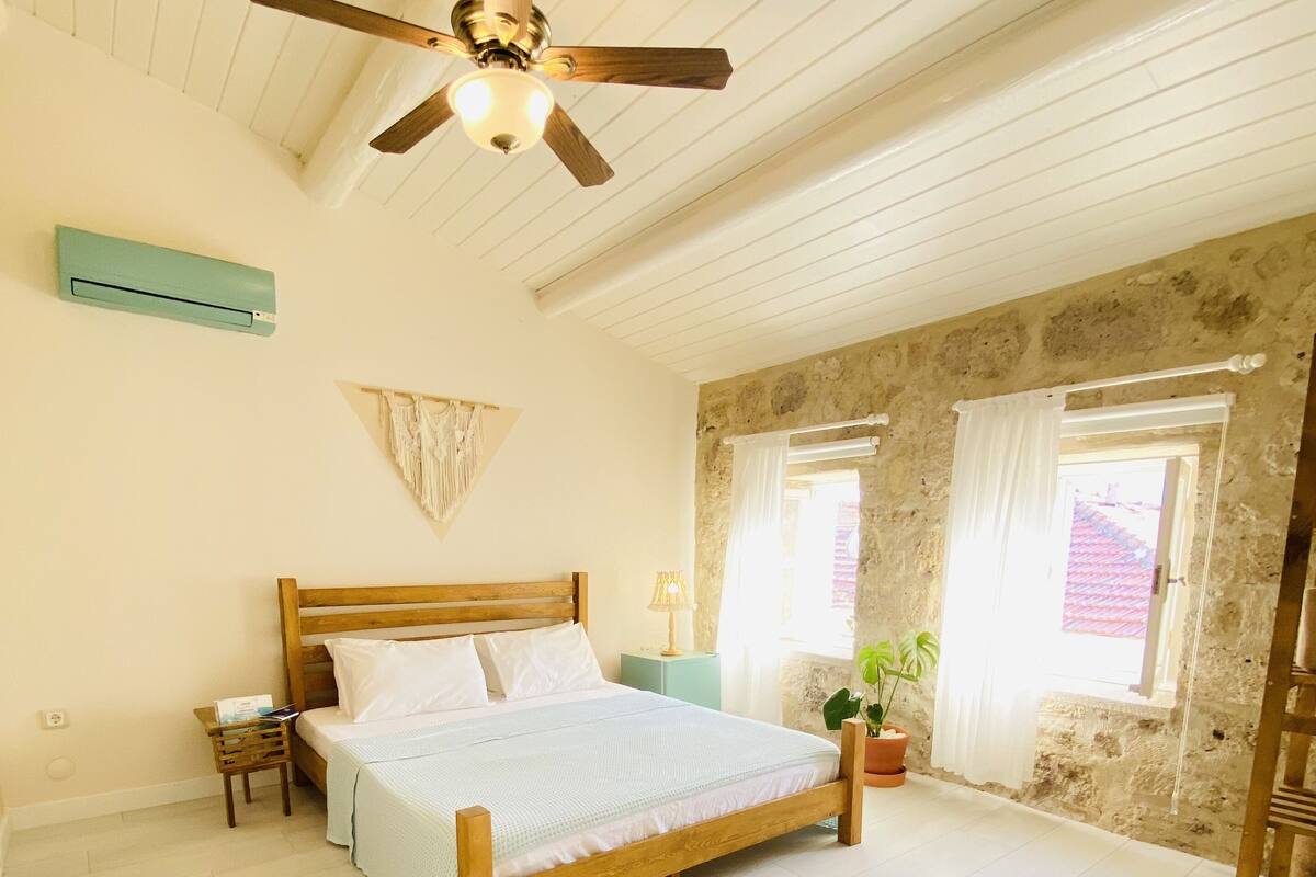 Un dormitorio con una cama de madera, un ventilador de techo, una mesita de noche pequeña, una planta en maceta y un sillón mullido.